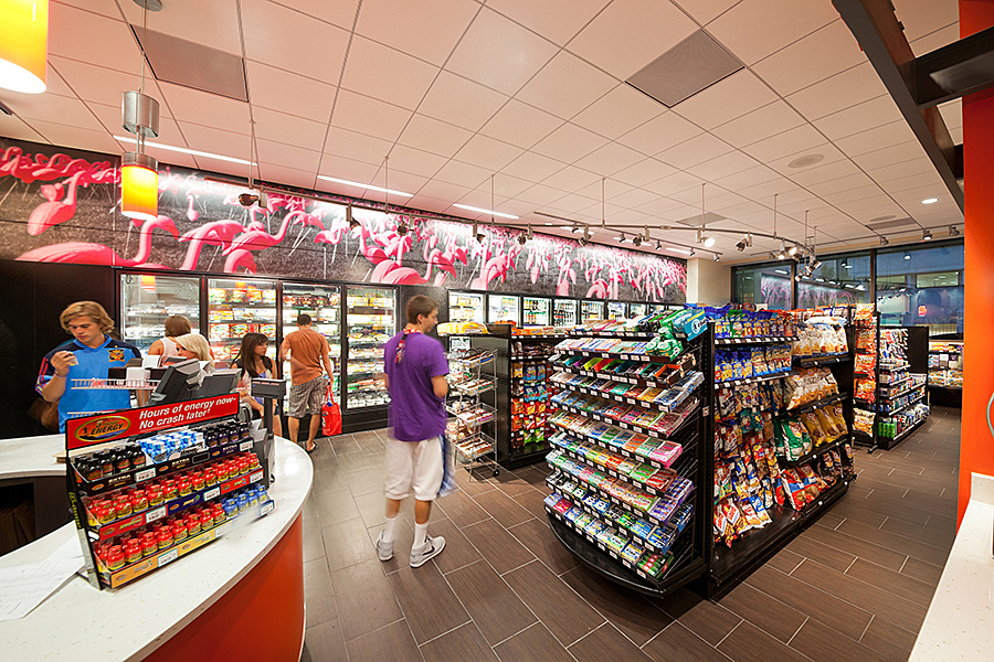 Decorative flamingo signage positioned above row of refrigerators while people browse for items at convenience store.