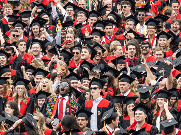 A joyful group of UW-Madison graduates in black caps and gowns with red accents celebrate together, smiling and raising their hands.