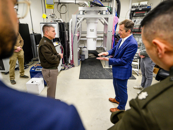A man in a blue suit speaks to another man in a military uniform. They surrounded by individuals in a laboratory setting.
