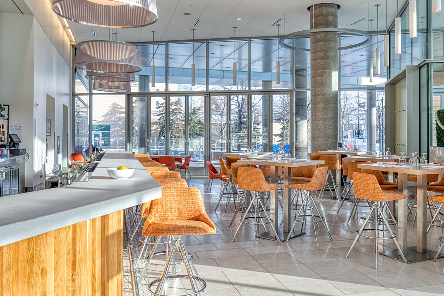 Bright dining area with bar, high-top tables and floor-to-ceiling glass windows during sunny day.